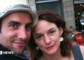 A close-up photograph shows a man and a woman - both with brown hair - posing together outdoors and looking toward the camera. The man wears a white hat. The backdrop is a city street with a building façade, outdoor seating and an orange scooter visible in the background.