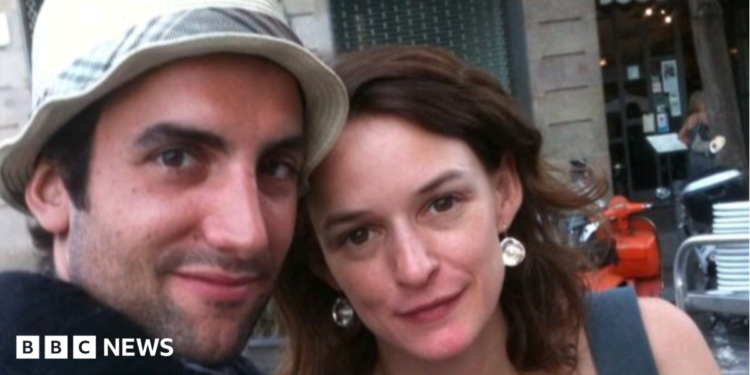 A close-up photograph shows a man and a woman - both with brown hair - posing together outdoors and looking toward the camera. The man wears a white hat. The backdrop is a city street with a building façade, outdoor seating and an orange scooter visible in the background.