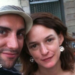 A close-up photograph shows a man and a woman - both with brown hair - posing together outdoors and looking toward the camera. The man wears a white hat. The backdrop is a city street with a building façade, outdoor seating and an orange scooter visible in the background.