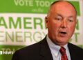 Pete Hoekstra, with short grey hair, and in a dark suit coat, red tie and patterned blue shirt, stands in from of a green and white campaign sign promoting American energy.
