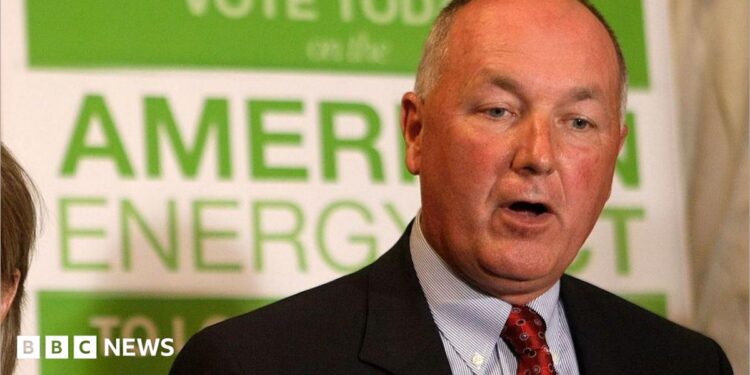 Pete Hoekstra, with short grey hair, and in a dark suit coat, red tie and patterned blue shirt, stands in from of a green and white campaign sign promoting American energy.