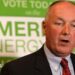 Pete Hoekstra, with short grey hair, and in a dark suit coat, red tie and patterned blue shirt, stands in from of a green and white campaign sign promoting American energy.