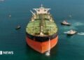 Stock image of an oil tanker pictured front on, with several tugs surrounding the ship
