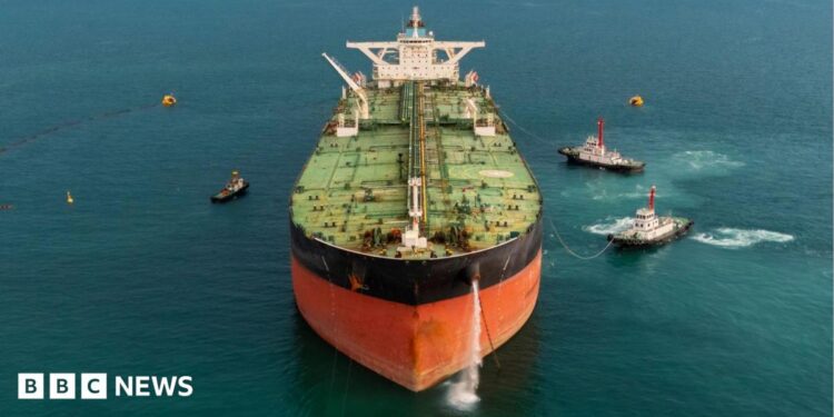Stock image of an oil tanker pictured front on, with several tugs surrounding the ship