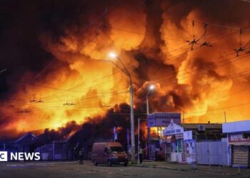 Large fire engulfs a market area at night, sending towering flames and thick smoke into the sky above nearby buildings, streets, and overhead power lines.