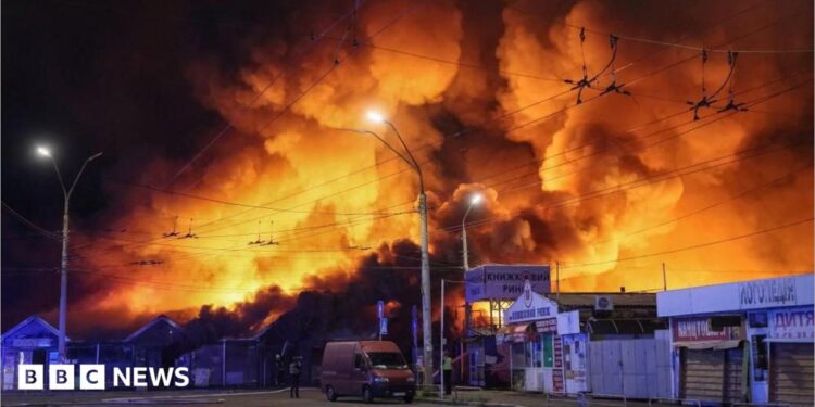 Large fire engulfs a market area at night, sending towering flames and thick smoke into the sky above nearby buildings, streets, and overhead power lines.