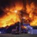 Large fire engulfs a market area at night, sending towering flames and thick smoke into the sky above nearby buildings, streets, and overhead power lines.