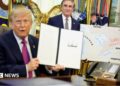 US President Donald Trump holds up a document with his signature in the Oval Office. Begin him is Secretary of the Interior Doug Burghum and a large map showing the US and Canada and the Great Lakes, with Lake Ontario renamed in red script as Lake America.
