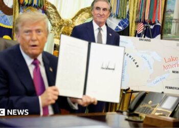 US President Donald Trump holds up a document with his signature in the Oval Office. Begin him is Secretary of the Interior Doug Burghum and a large map showing the US and Canada and the Great Lakes, with Lake Ontario renamed in red script as Lake America.