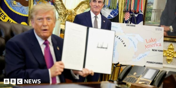 US President Donald Trump holds up a document with his signature in the Oval Office. Begin him is Secretary of the Interior Doug Burghum and a large map showing the US and Canada and the Great Lakes, with Lake Ontario renamed in red script as Lake America.