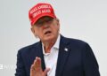 A close up shot of President Donald Trump speaking to outside with his hand raised. He is wearing a "make America great again" cap