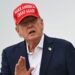 A close up shot of President Donald Trump speaking to outside with his hand raised. He is wearing a "make America great again" cap