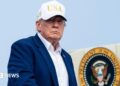 Trump, wearing a suit and no tie and a white "USA" hat, descending from airplane steps