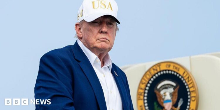 Trump, wearing a suit and no tie and a white "USA" hat, descending from airplane steps