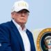 Trump, wearing a suit and no tie and a white "USA" hat, descending from airplane steps