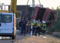 A crane winches an overturned bus up on the side of the road as firefighters look on.