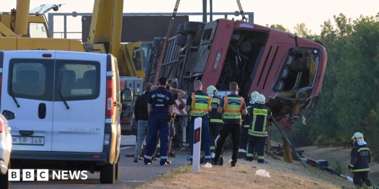 A crane winches an overturned bus up on the side of the road as firefighters look on.