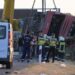 A crane winches an overturned bus up on the side of the road as firefighters look on.