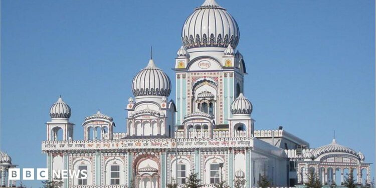 An image showing the outside of the Nanaksar Gurdwara Gursikh Temple Edmonton. It is a large, ornate white building with baby blue and rose colourings on its facade, as well as arches and several domed roofs. It is seen surrounded by spruce trees.