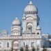 An image showing the outside of the Nanaksar Gurdwara Gursikh Temple Edmonton. It is a large, ornate white building with baby blue and rose colourings on its facade, as well as arches and several domed roofs. It is seen surrounded by spruce trees.