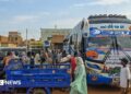 A coach is parked on the right-hand side of the picture. A group of people are standing close by gathering luggage. In the foreground, an open-backed tuktuk is parked.