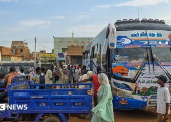 A coach is parked on the right-hand side of the picture. A group of people are standing close by gathering luggage. In the foreground, an open-backed tuktuk is parked.