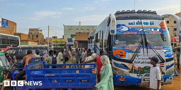 A coach is parked on the right-hand side of the picture. A group of people are standing close by gathering luggage. In the foreground, an open-backed tuktuk is parked.
