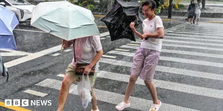 People holding umbrellas along a street in Shanghai. One boy is seen trying to hold on to an inverted umbrella as winds intensify
