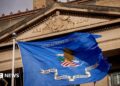 Close up of a DOJ flag (a rectangular base background of ultramarine blue, bearing an eagle on a shield, a scroll and the inscription 'Department of Justice.' The eagle faces to the left, with its left claw holding 13 arrows with the tips facing down. Its right claw holds an olive branch. The shield consists of a white base, a blue chief and six scarlet stripes. The scroll shall read in bold blue letters, 'QUI PRO DOMINA JUSTITIA SEQUITUR' - meaning, Who for Lady Justice prosecutes) flying in front of the DOJ headquarters