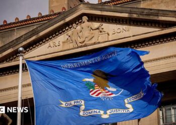 Close up of a DOJ flag (a rectangular base background of ultramarine blue, bearing an eagle on a shield, a scroll and the inscription 'Department of Justice.' The eagle faces to the left, with its left claw holding 13 arrows with the tips facing down. Its right claw holds an olive branch. The shield consists of a white base, a blue chief and six scarlet stripes. The scroll shall read in bold blue letters, 'QUI PRO DOMINA JUSTITIA SEQUITUR' - meaning, Who for Lady Justice prosecutes) flying in front of the DOJ headquarters
