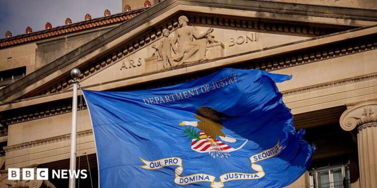 Close up of a DOJ flag (a rectangular base background of ultramarine blue, bearing an eagle on a shield, a scroll and the inscription 'Department of Justice.' The eagle faces to the left, with its left claw holding 13 arrows with the tips facing down. Its right claw holds an olive branch. The shield consists of a white base, a blue chief and six scarlet stripes. The scroll shall read in bold blue letters, 'QUI PRO DOMINA JUSTITIA SEQUITUR' - meaning, Who for Lady Justice prosecutes) flying in front of the DOJ headquarters