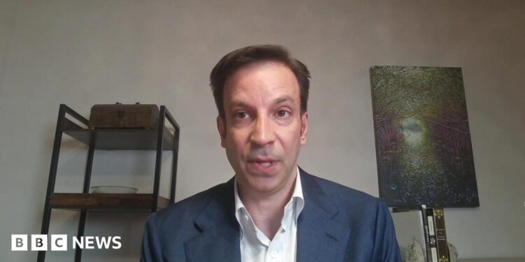 Erik Slavin wearing a white shirt and blue blazer, a grey wall in the background with dark metal-framed shelves with objects on display to his right, and a textured, greenish relief object on a thin stand to his left.