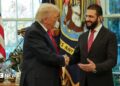 Two men in dark suits and red ties smile and shake hands in front of windows and American flags