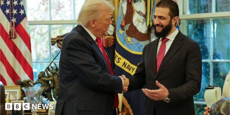 Two men in dark suits and red ties smile and shake hands in front of windows and American flags