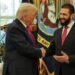 Two men in dark suits and red ties smile and shake hands in front of windows and American flags