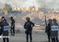Spanish armed police with riot shields on Trampolin beach, Ceuta, as anti-migrant protesters set fire to a migrant camp there. Their faces are blurred. Smoke can be seen rising in the air.