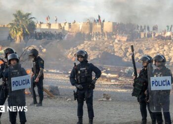 Spanish armed police with riot shields on Trampolin beach, Ceuta, as anti-migrant protesters set fire to a migrant camp there. Their faces are blurred. Smoke can be seen rising in the air.