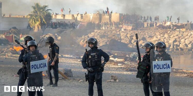Spanish armed police with riot shields on Trampolin beach, Ceuta, as anti-migrant protesters set fire to a migrant camp there. Their faces are blurred. Smoke can be seen rising in the air.