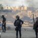 Spanish armed police with riot shields on Trampolin beach, Ceuta, as anti-migrant protesters set fire to a migrant camp there. Their faces are blurred. Smoke can be seen rising in the air.
