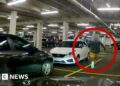 A man in blue sneakers is circled in red, running through a car park.