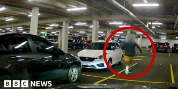 A man in blue sneakers is circled in red, running through a car park.