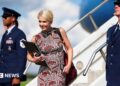 Natalie Harp walks off Air Force One wearing a sleeveless printed red dress. She is smiling and has a laptop in her hands.