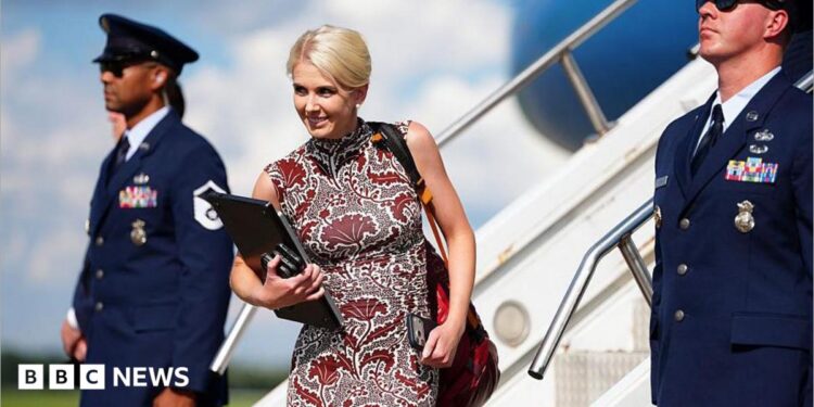 Natalie Harp walks off Air Force One wearing a sleeveless printed red dress. She is smiling and has a laptop in her hands.