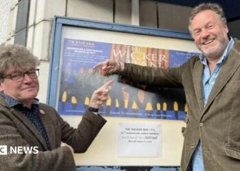 Two men in their 50s either side of a poster for The Wicker Man outside a cinema. Dominic, on the left, is in a tweed suit and has glasses and thick hair. Justin (right) is in a cord suit and has a beard. They are smiling and pointing at the poster.