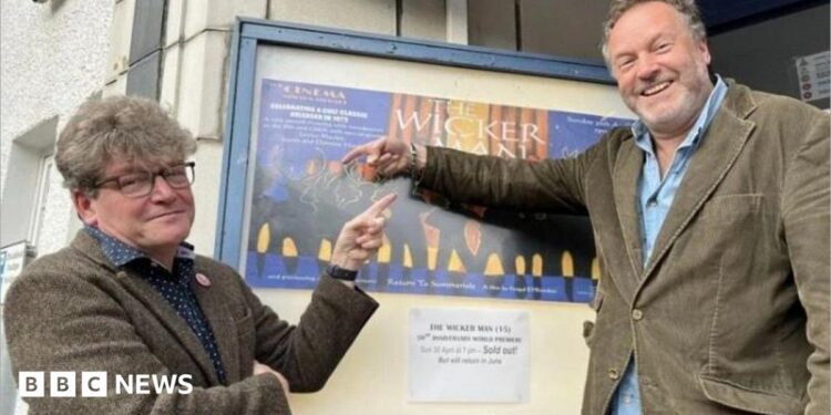 Two men in their 50s either side of a poster for The Wicker Man outside a cinema. Dominic, on the left, is in a tweed suit and has glasses and thick hair. Justin (right) is in a cord suit and has a beard. They are smiling and pointing at the poster.
