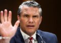 Pete Hegseth gestures by lifting his hand as he frowns