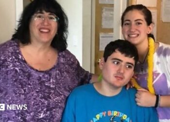 Linda Mancuso, wearing a purple top, glasses and dark hair falling to her shoulders, stands next to her 23-year-old son, Michael, who needs 24/7 care; he wears a "Happy Birthday" blue shirt and stares off to the side; her daughter stands to the other side of Michael, smiling as she rests a hand on his shoulder
