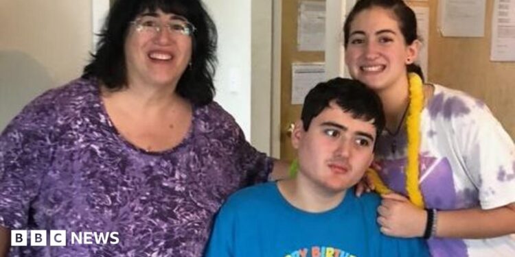 Linda Mancuso, wearing a purple top, glasses and dark hair falling to her shoulders, stands next to her 23-year-old son, Michael, who needs 24/7 care; he wears a "Happy Birthday" blue shirt and stares off to the side; her daughter stands to the other side of Michael, smiling as she rests a hand on his shoulder