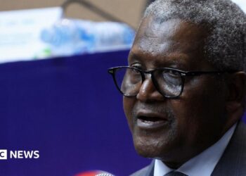A close-up photo of Aliko Dangote. He is wearing glasses and a suit, with short, salt and pepper hair.