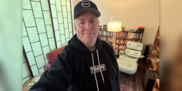 Andrew Wimsatt in a selfie photo taken inside a home library while wearing a black hoodie and ballcap